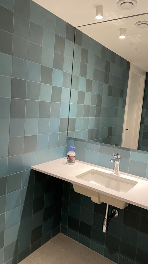Baño de oficina con pared de azulejos verdes y espejo amplio